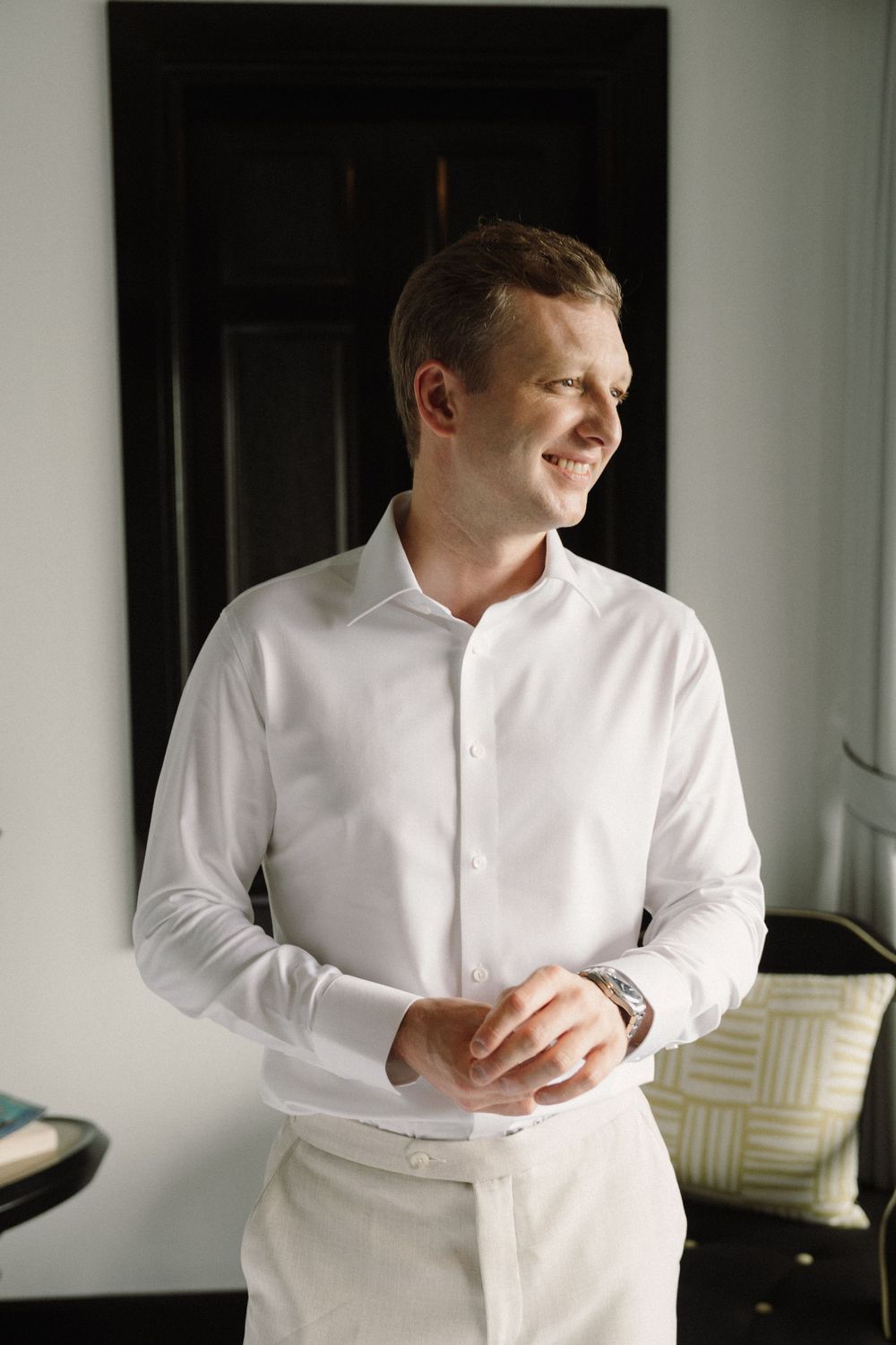 A groom in a crisp white dress shirt adjusts his attire while getting ready for the wedding.