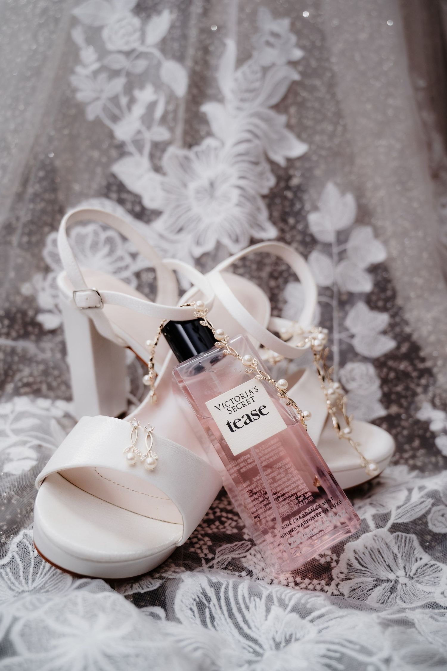 White lace bridal shoes and perfume bottle arranged on delicate lace fabric with soft pink accents.