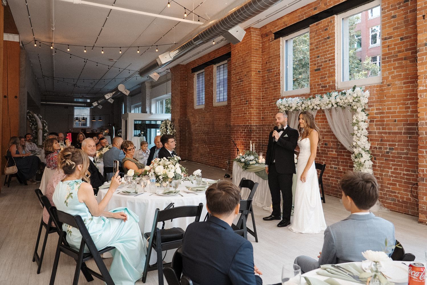 Romantic wedding reception table setting with candlesticks and floral centerpieces against exposed brick wall and flowing drapes.