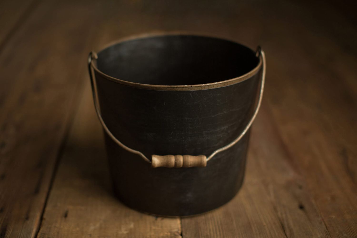 A rustic black metal bucket with wooden handle sits on a dark wooden surface in moody lighting.