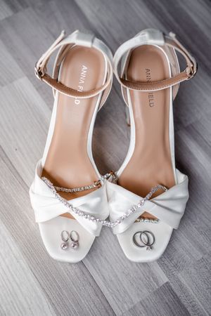 Elegant white bridal shoes with crystal embellishments and wedding rings displayed on gray floor.