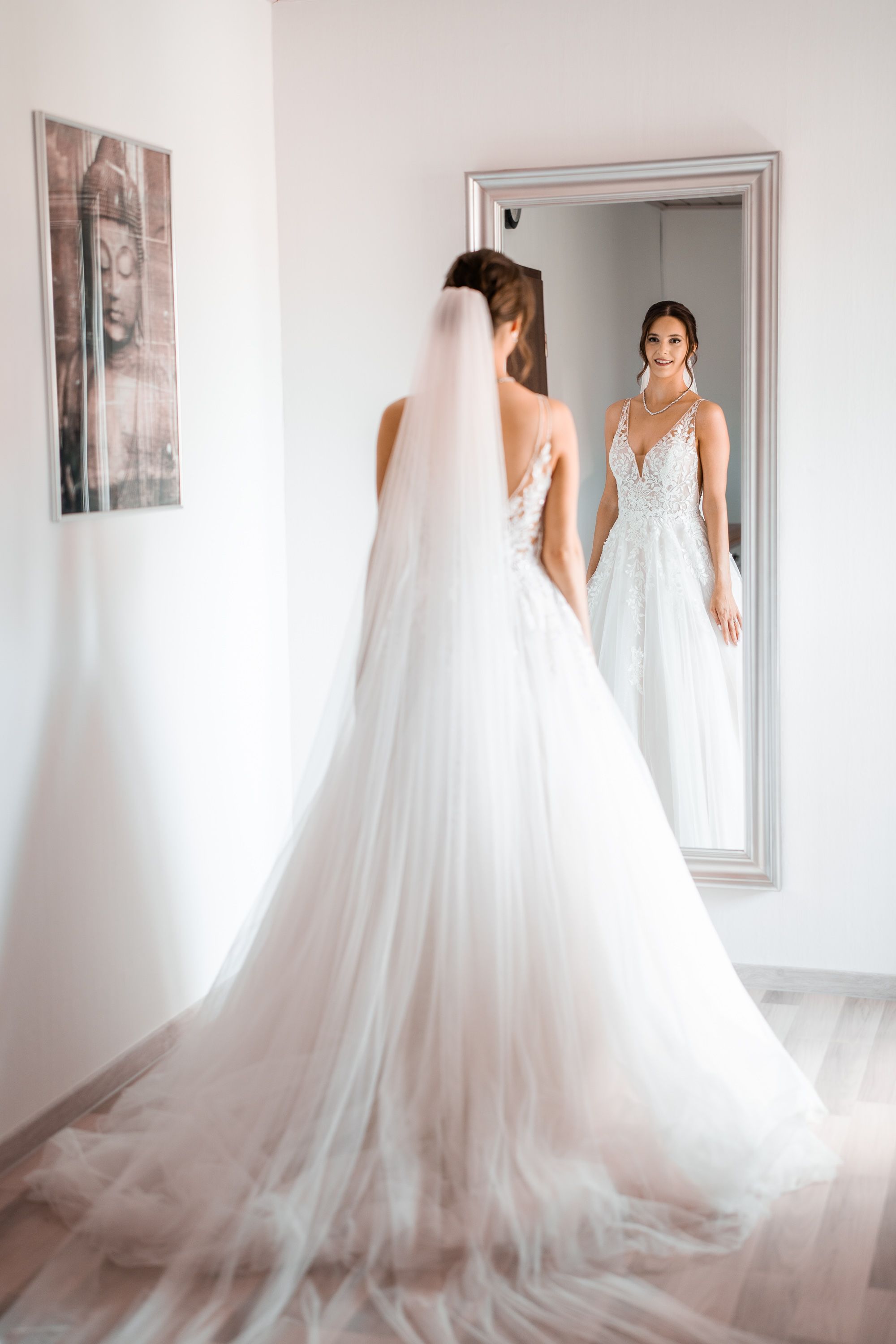 Elegant wedding dress with flowing tulle train reflected in a full-length mirror in a bright white room.