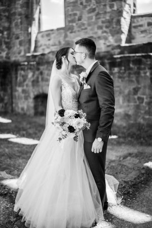 Black and white wedding portrait of couple with bouquet against stone wall in sunlight.