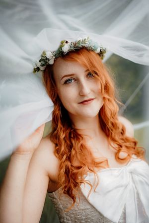 Ethereal bridal portrait with flowing copper hair and white floral crown against soft draped fabric background.