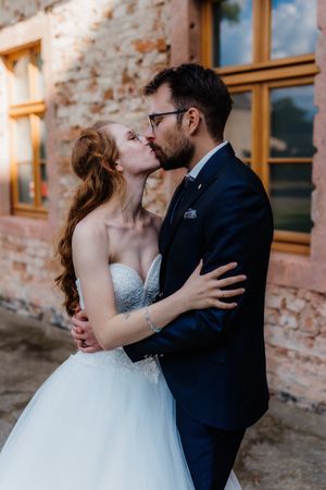 Romantic wedding kiss between couple in front of rustic brick building with large windows.