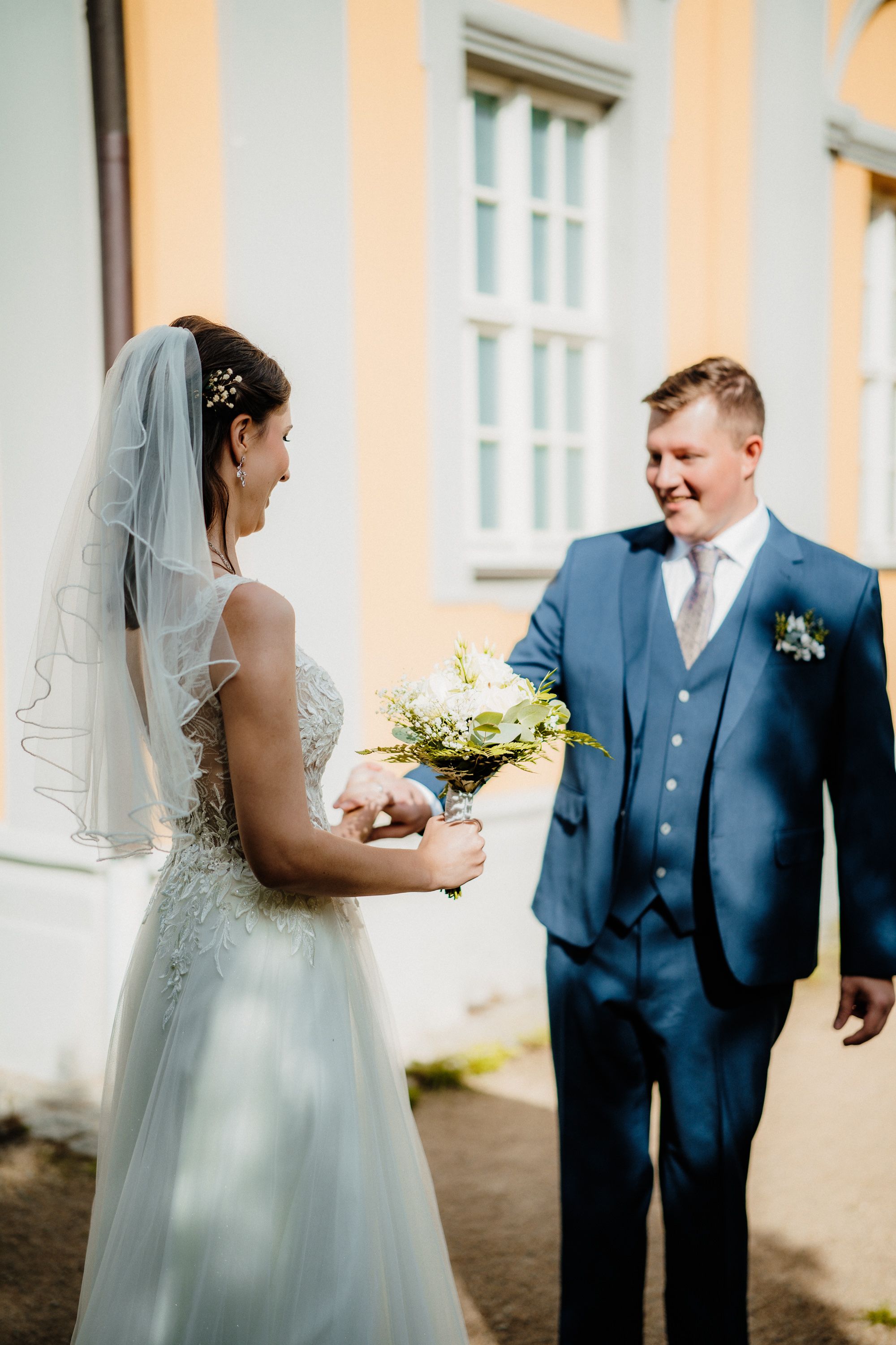 A romantic moment between newlyweds sharing glances and smiles outside a building with peach-colored walls.