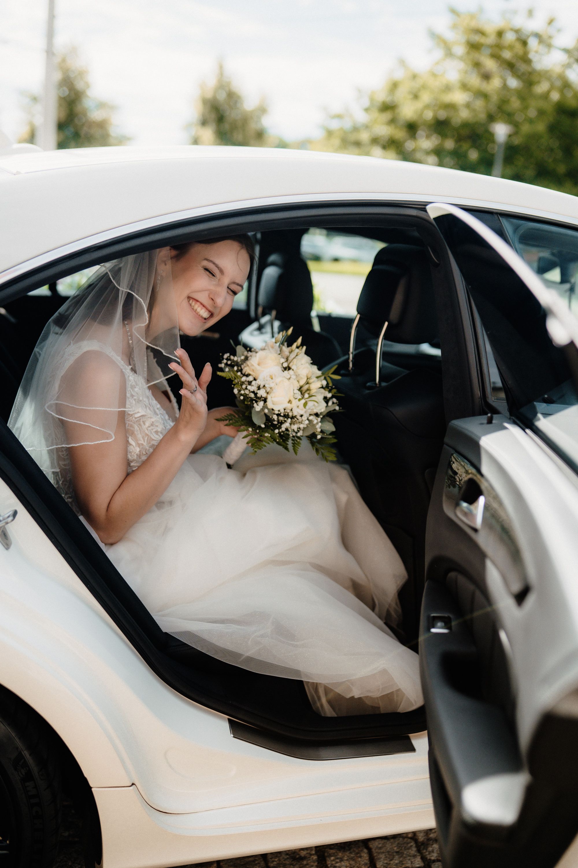 Laughing bride sitting in the back seat of a wedding car holding a bouquet of white flowers.