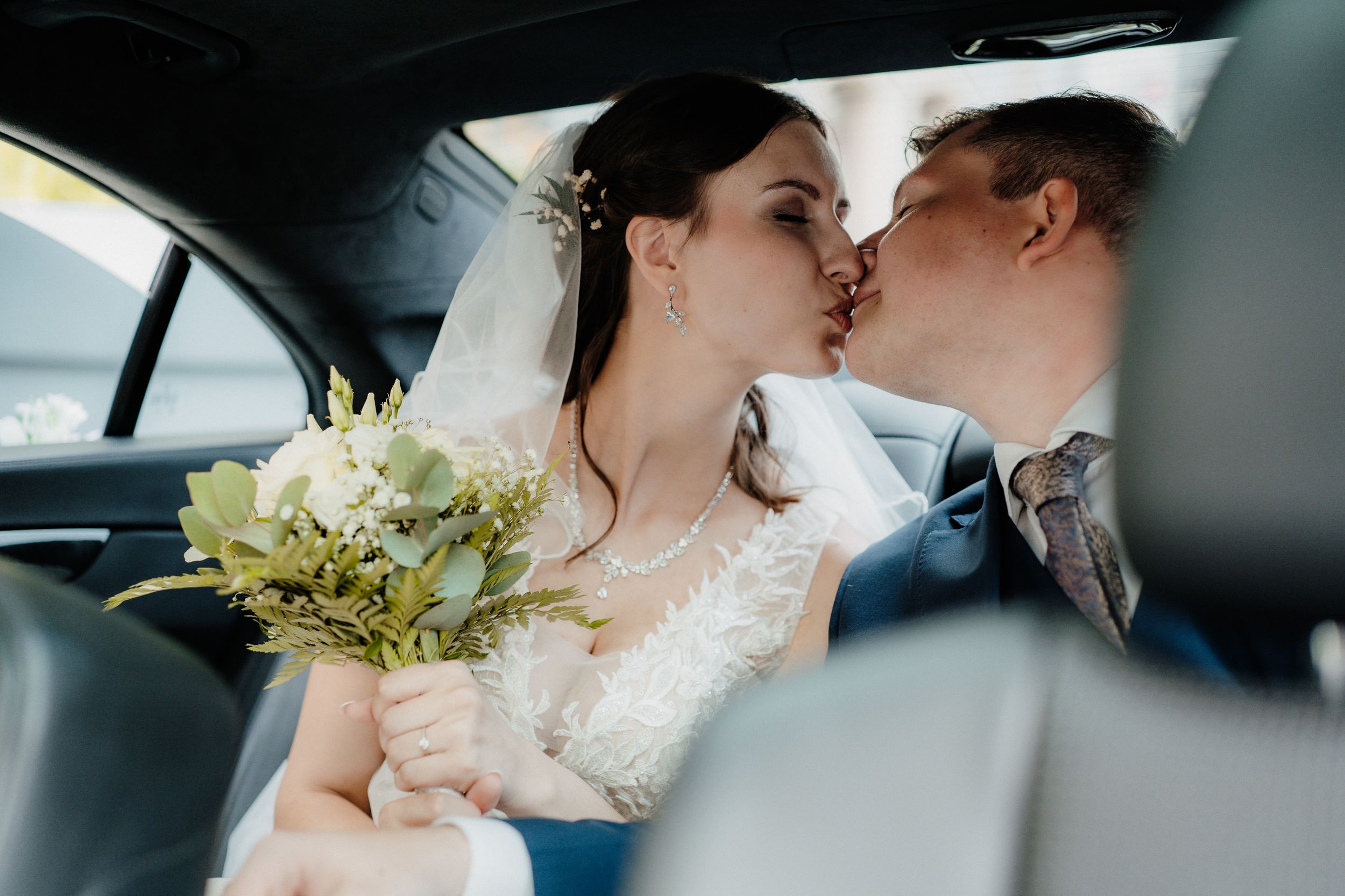 Newlyweds share a tender kiss in a car while the bride holds an elegant white and green floral bouquet.