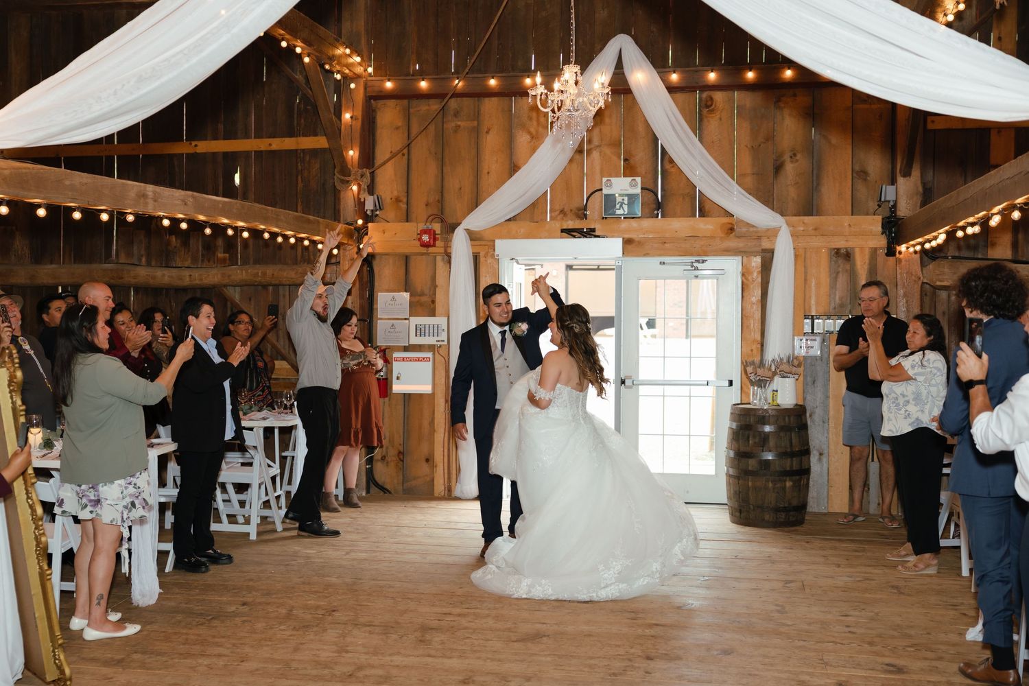 Couples dancing in a rustic, elegantly decorated barn venue with white fabric draping and string lights along the ceiling.