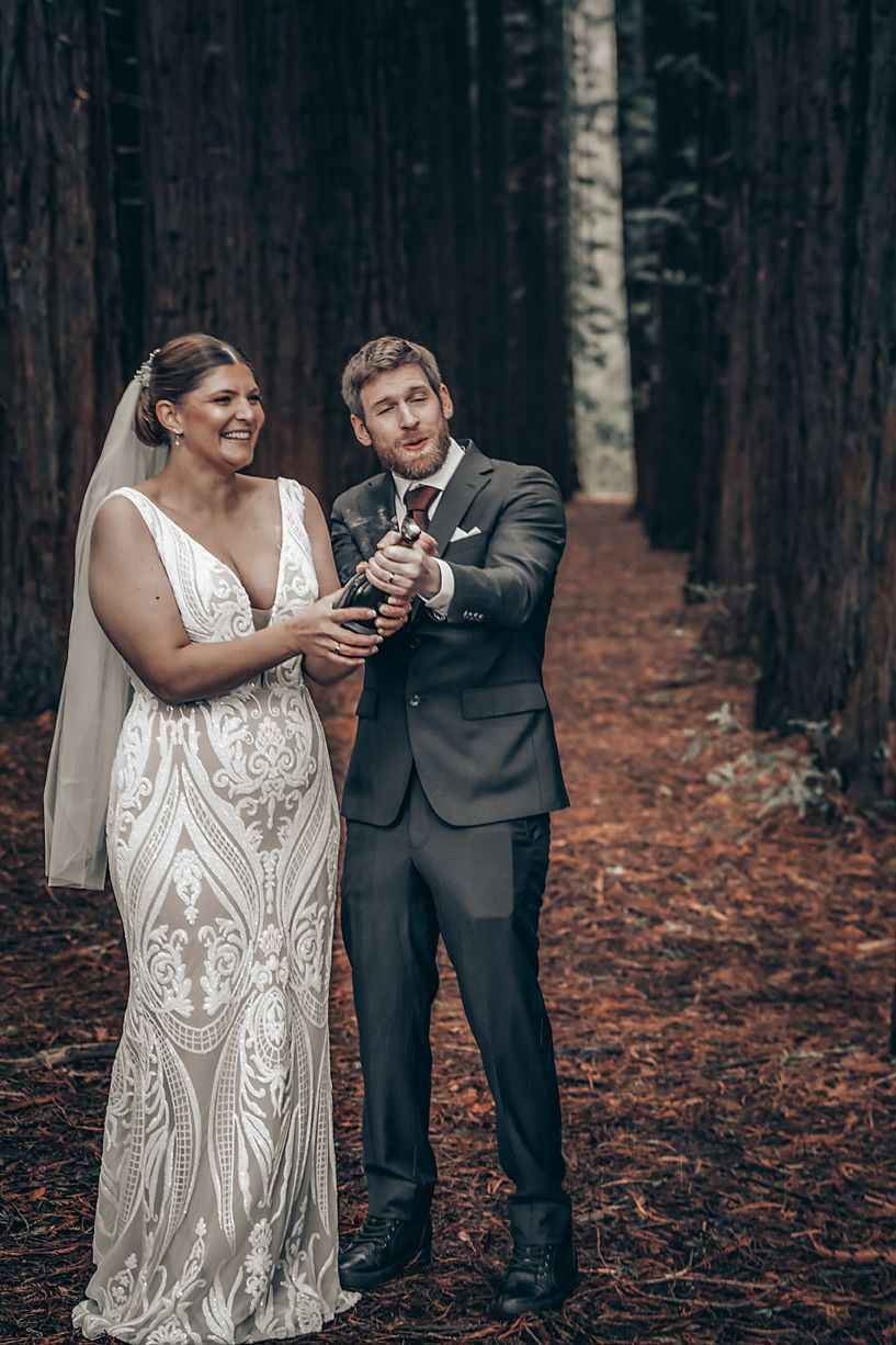 A bride in a white lace gown and groom in dark suit share a playful moment checking phones in a misty forest setting.