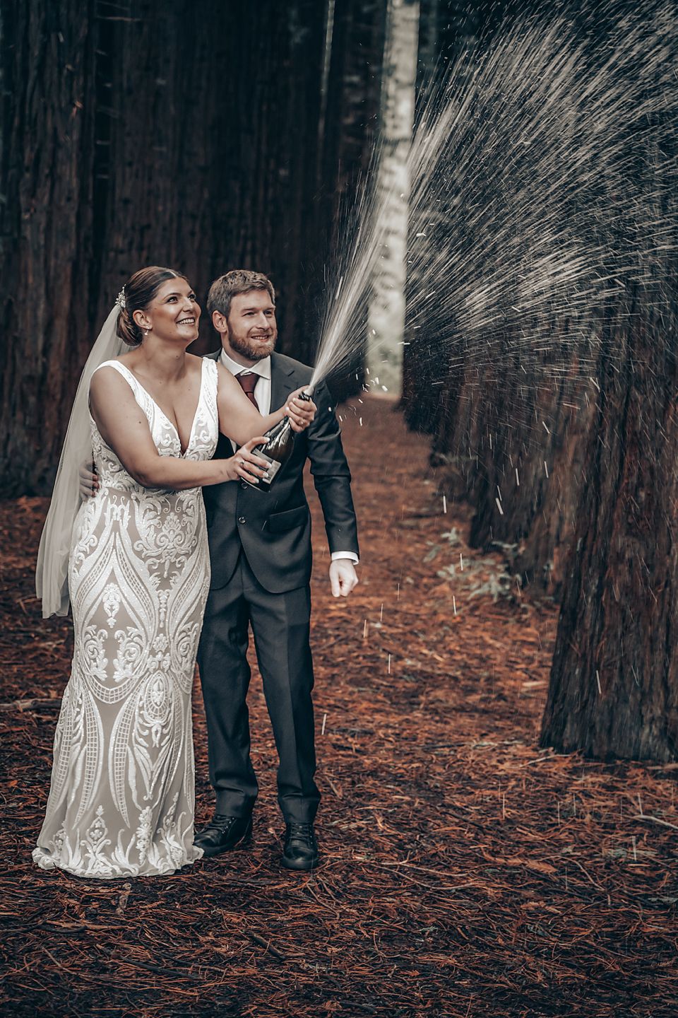 A couple in wedding attire take playful photos in a moody forest setting with fallen leaves beneath tall trees.