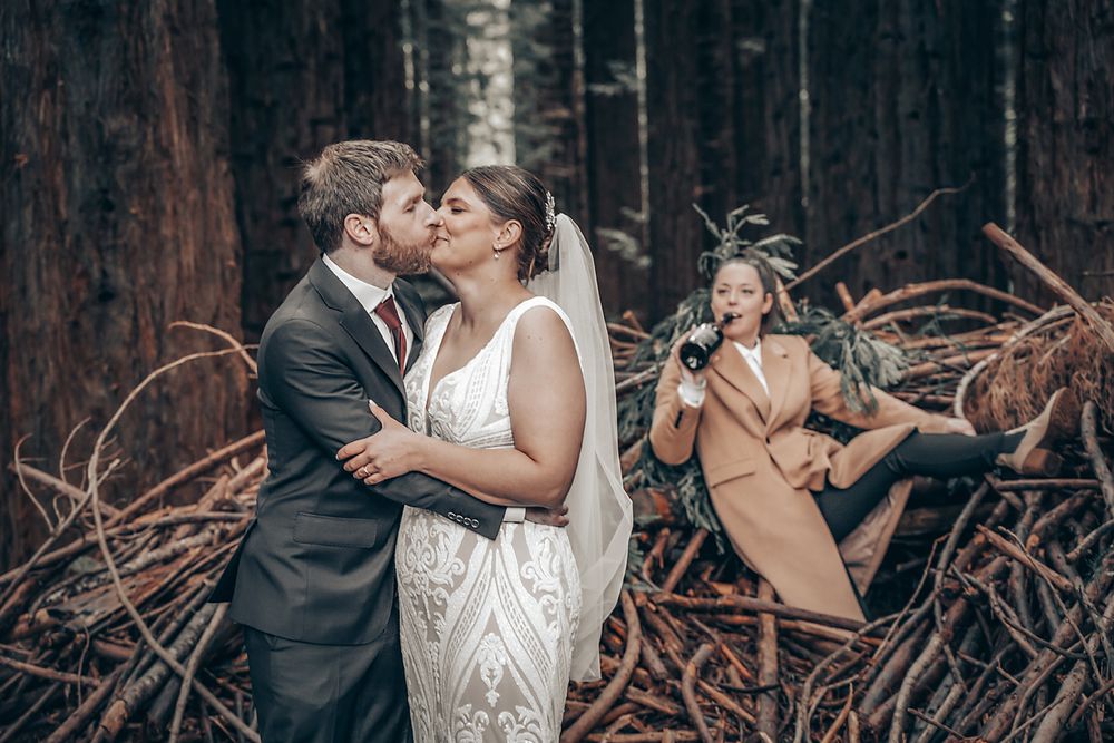 A couple shares a romantic kiss in a forest while a person in a tan coat poses playfully in the background.