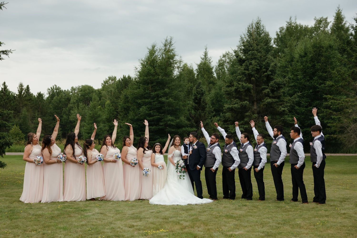 Wedding party poses in a creative formation on a green lawn with evergreen trees in the background.