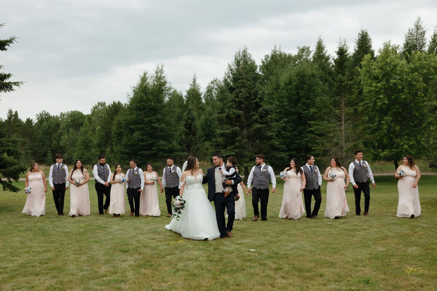 Wedding party in formal attire stands in line formation on a grassy field with evergreen trees in the background.