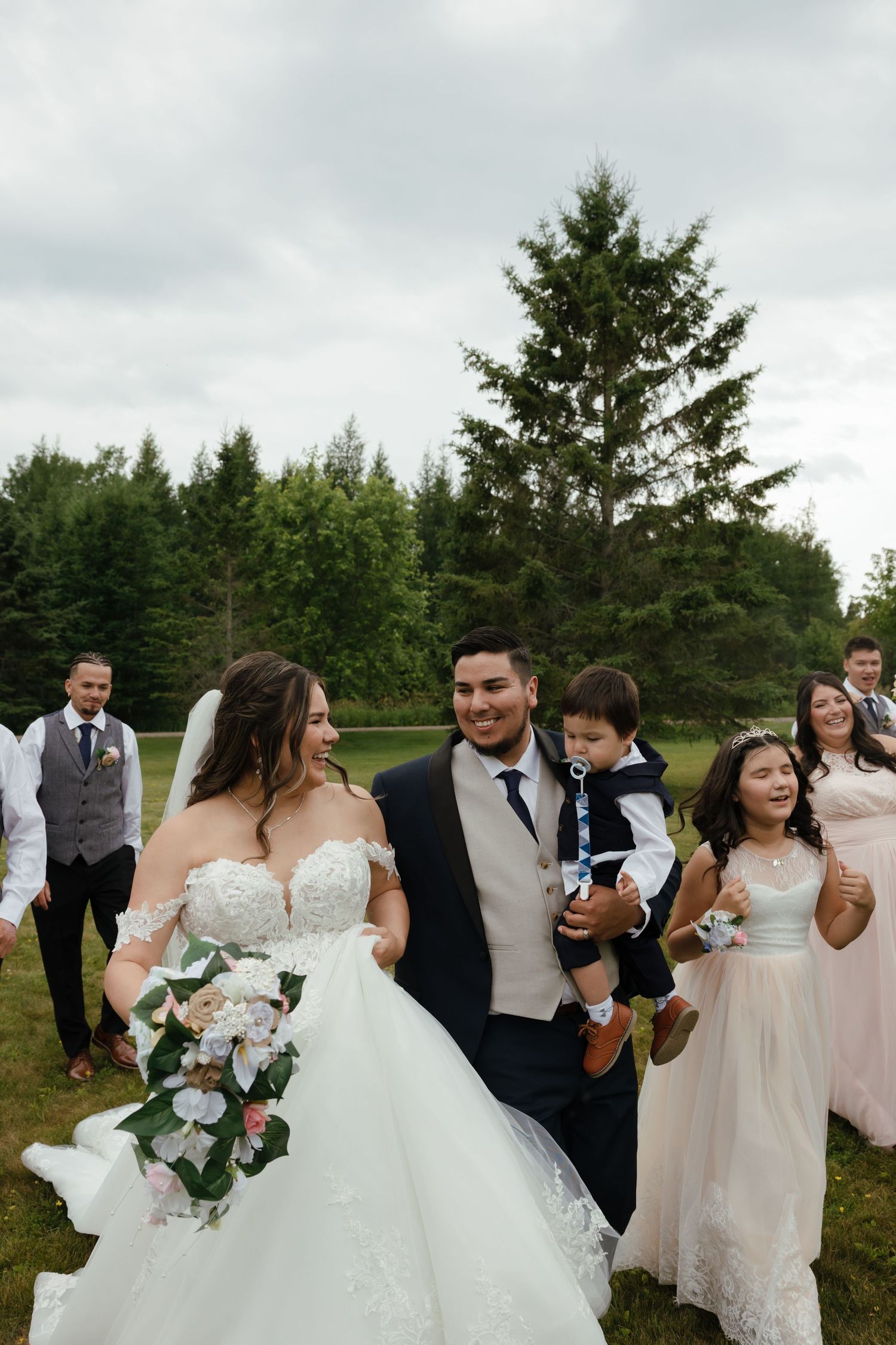 Wedding celebration on a lush lawn surrounded by tall evergreen trees on an overcast day.