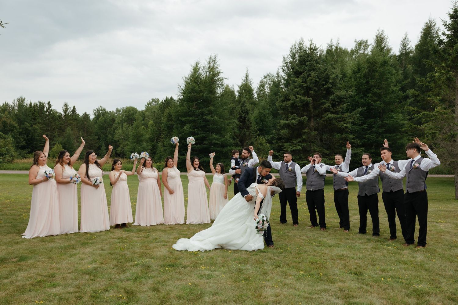 Wedding party members lined up in a grassy field wearing light pink dresses and dark suits pose for a group photo.