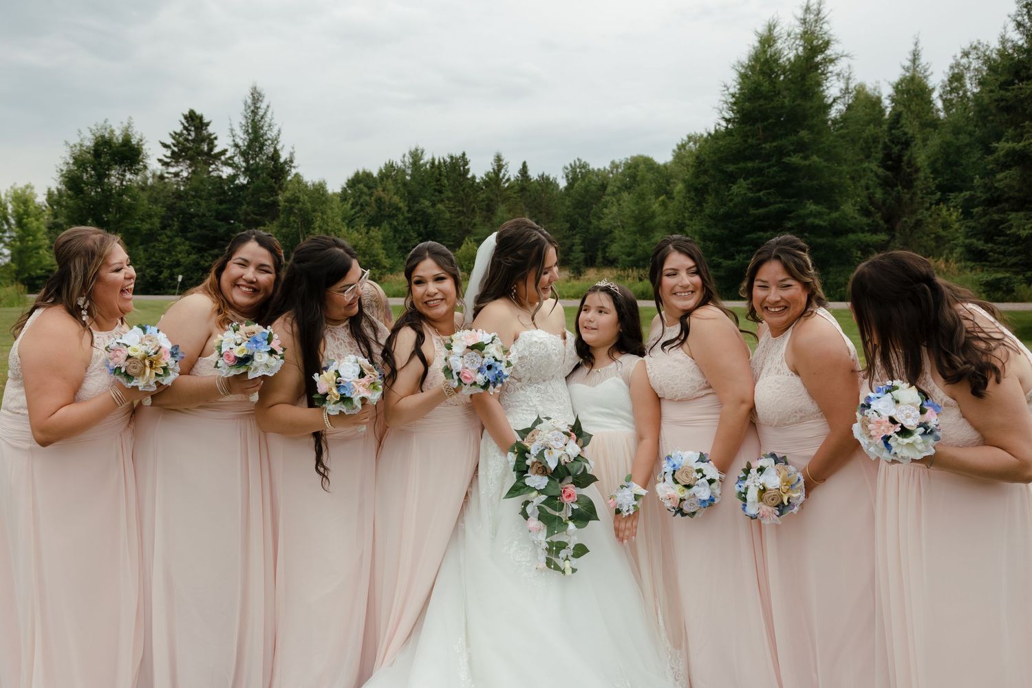 Bridesmaids in blush pink dresses stand together at an outdoor wedding celebration with a wooded backdrop.