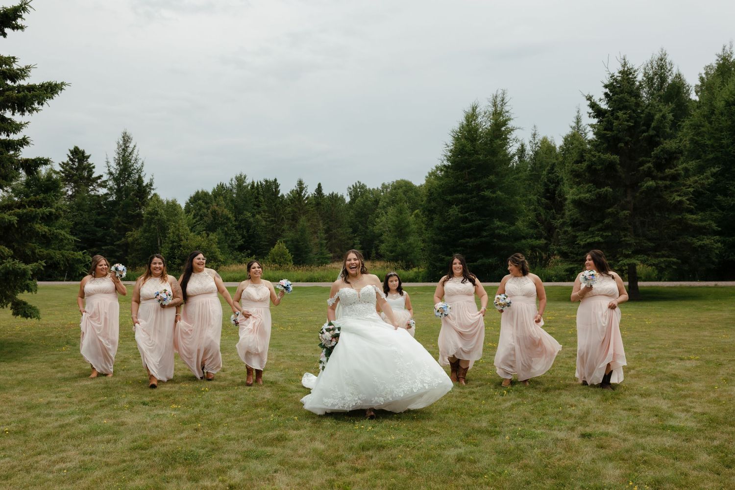 Bridesmaids in pink dresses dancing and celebrating with bride on grassy field with pine trees.