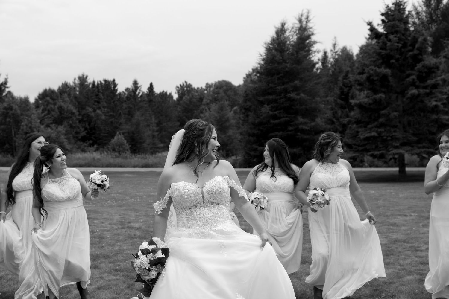A group in flowing white dresses stands together in a field with trees while posing for a panoramic photo series.