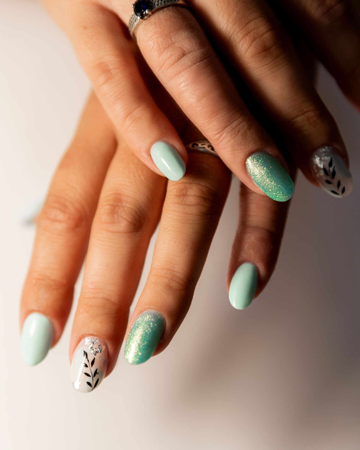 Close-up of mint green manicured nails with glitter accent and delicate black branch designs on selected nails.