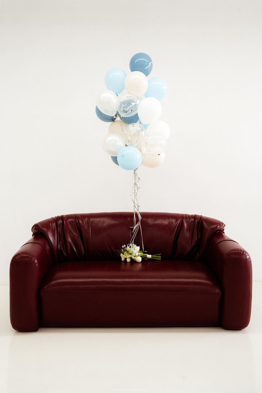 Blue and white balloons tied to bouquet floating above dark red leather sofa in studio.