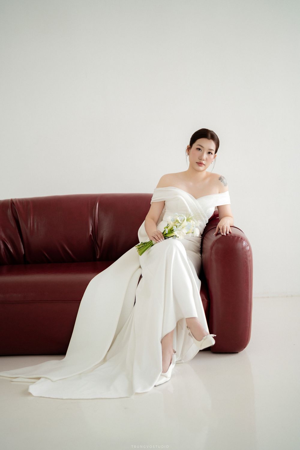 Elegant bride in white off-shoulder gown with train seated on dark red leather sofa.