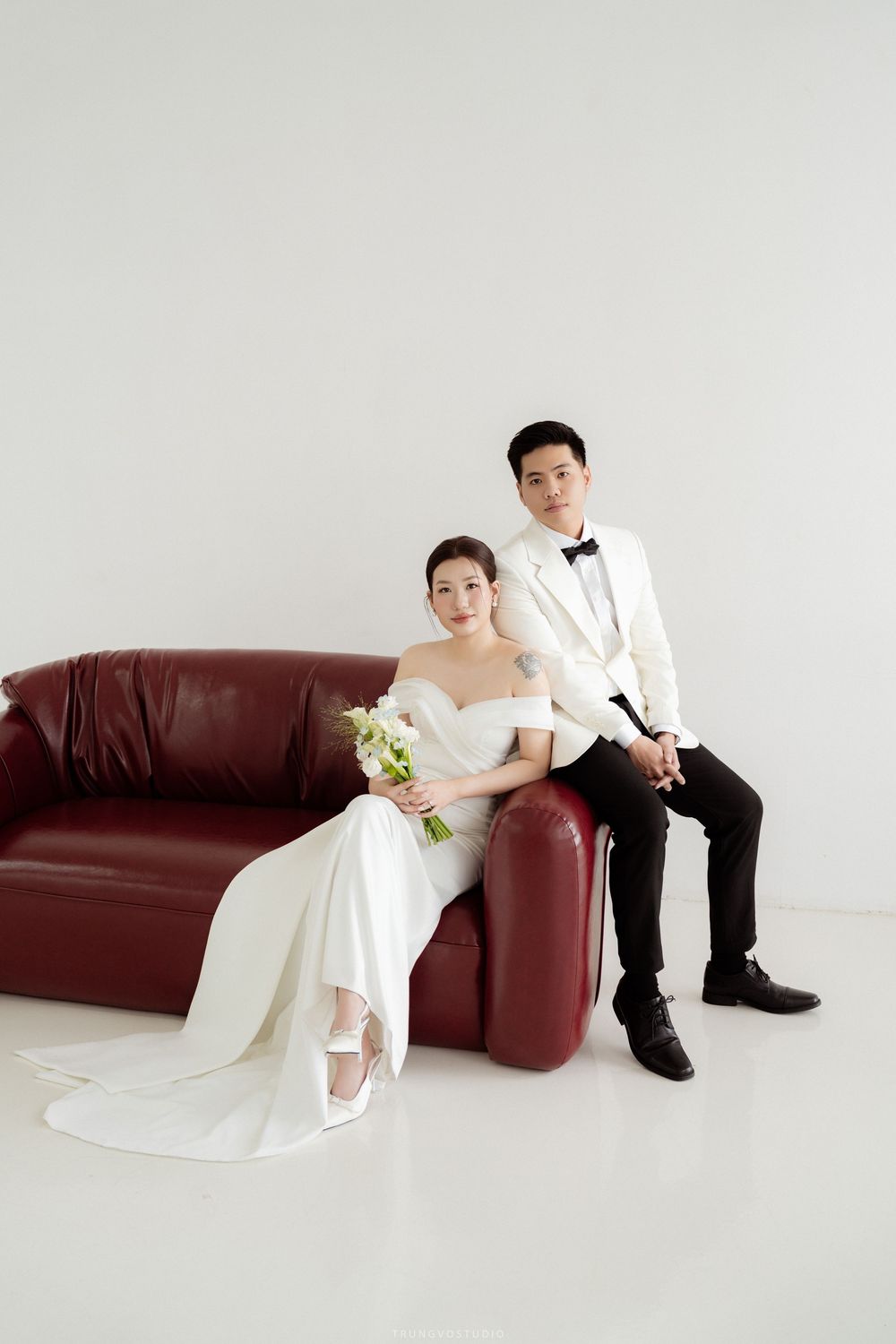 Bride and groom posing together on red leather sofa in white studio wedding portrait.