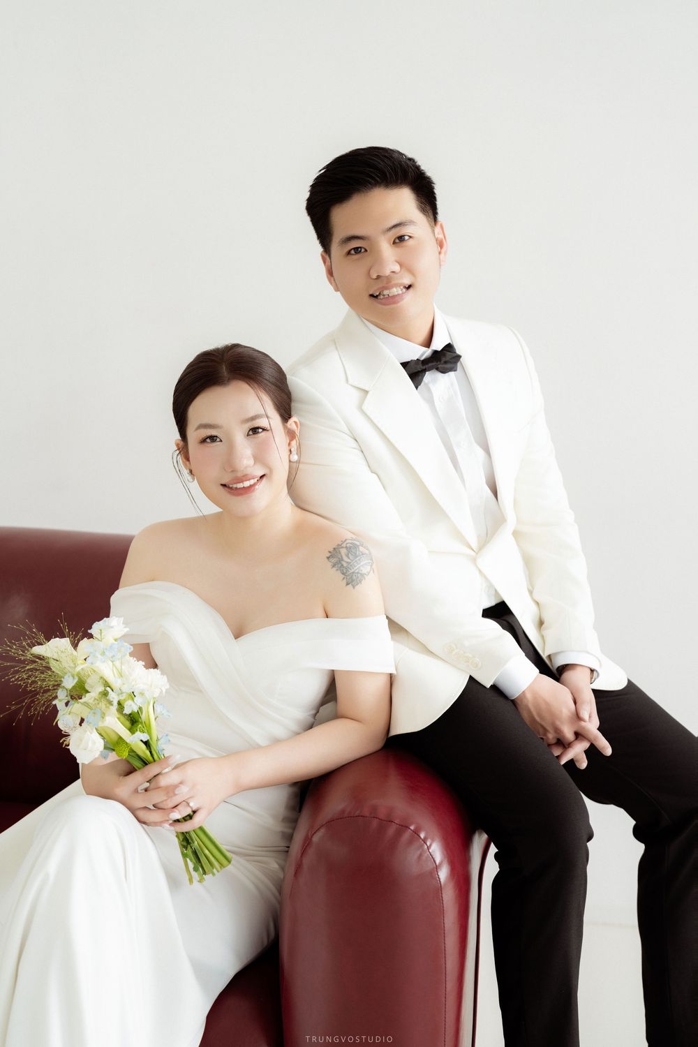 Smiling bride and groom in white tuxedo posing on red sofa in bright studio setting.