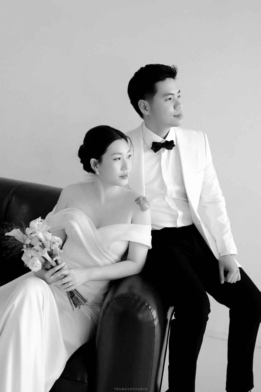 Black and white wedding photo of bride in off-shoulder gown with bouquet and groom in white tuxedo on sofa.