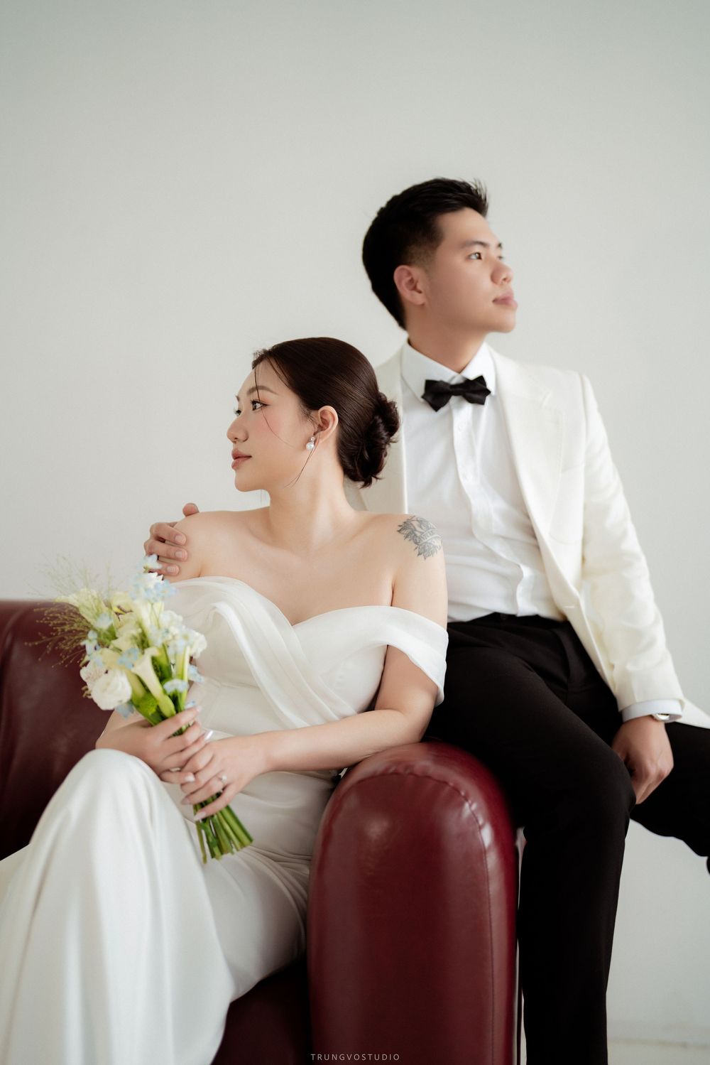 Elegant couple in wedding attire sitting on red leather sofa, bride holding white floral bouquet in studio.