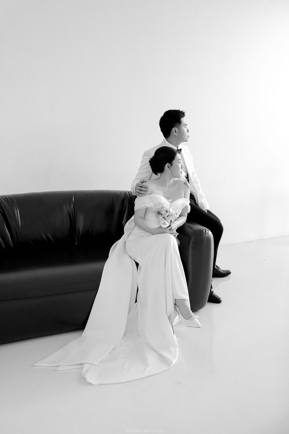 Black and white full-length wedding portrait of bride embracing groom seated on leather sofa in studio.