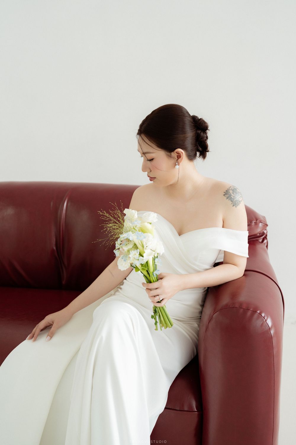 Bride in off-shoulder white gown sitting on red leather sofa holding white flower bouquet, looking down.