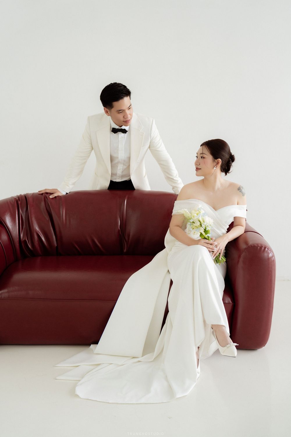 Wedding couple on red leather sofa, groom in white tuxedo standing behind seated bride with bouquet.