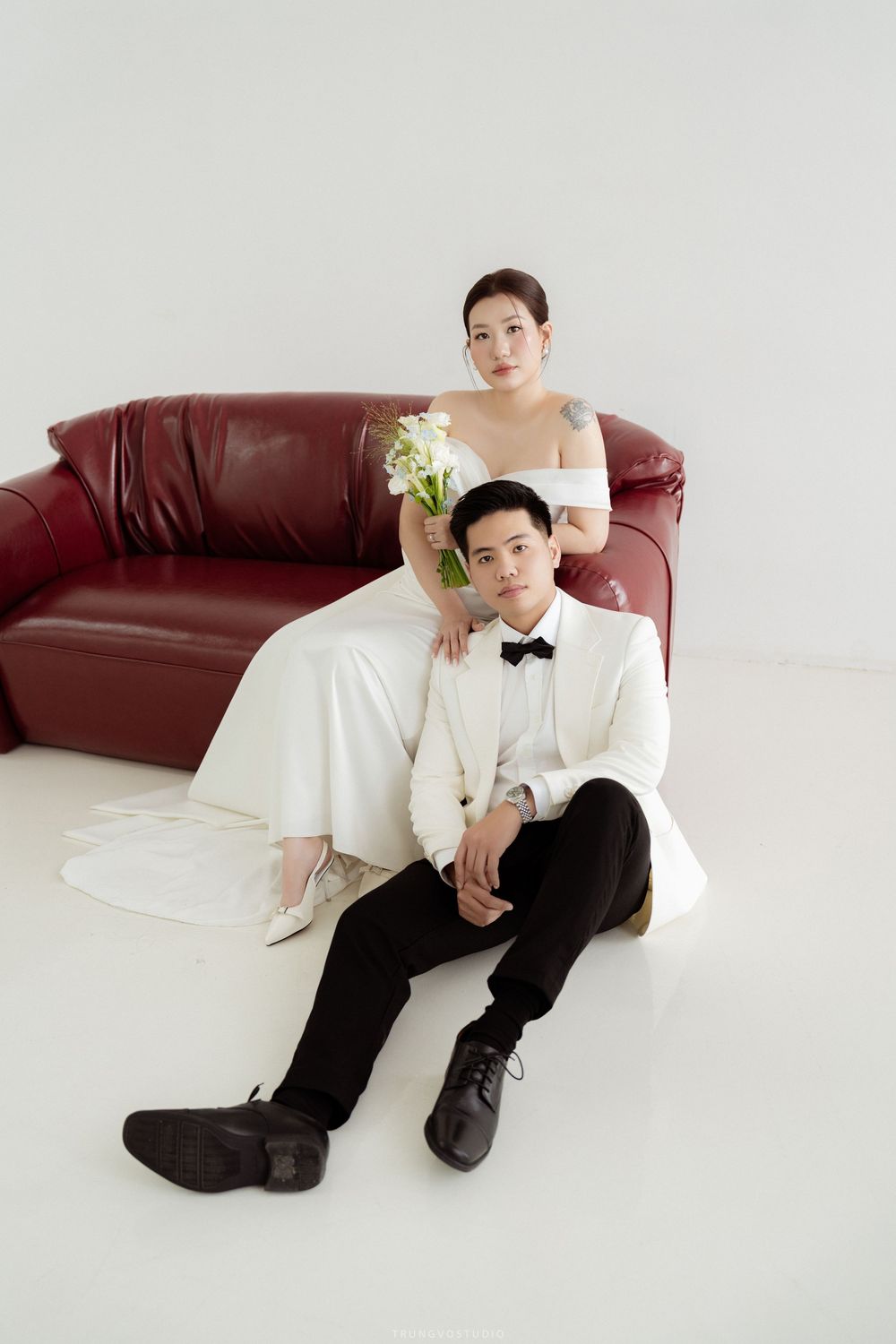 Groom in white tuxedo sits on floor leaning against red sofa while bride poses behind him with bouquet.