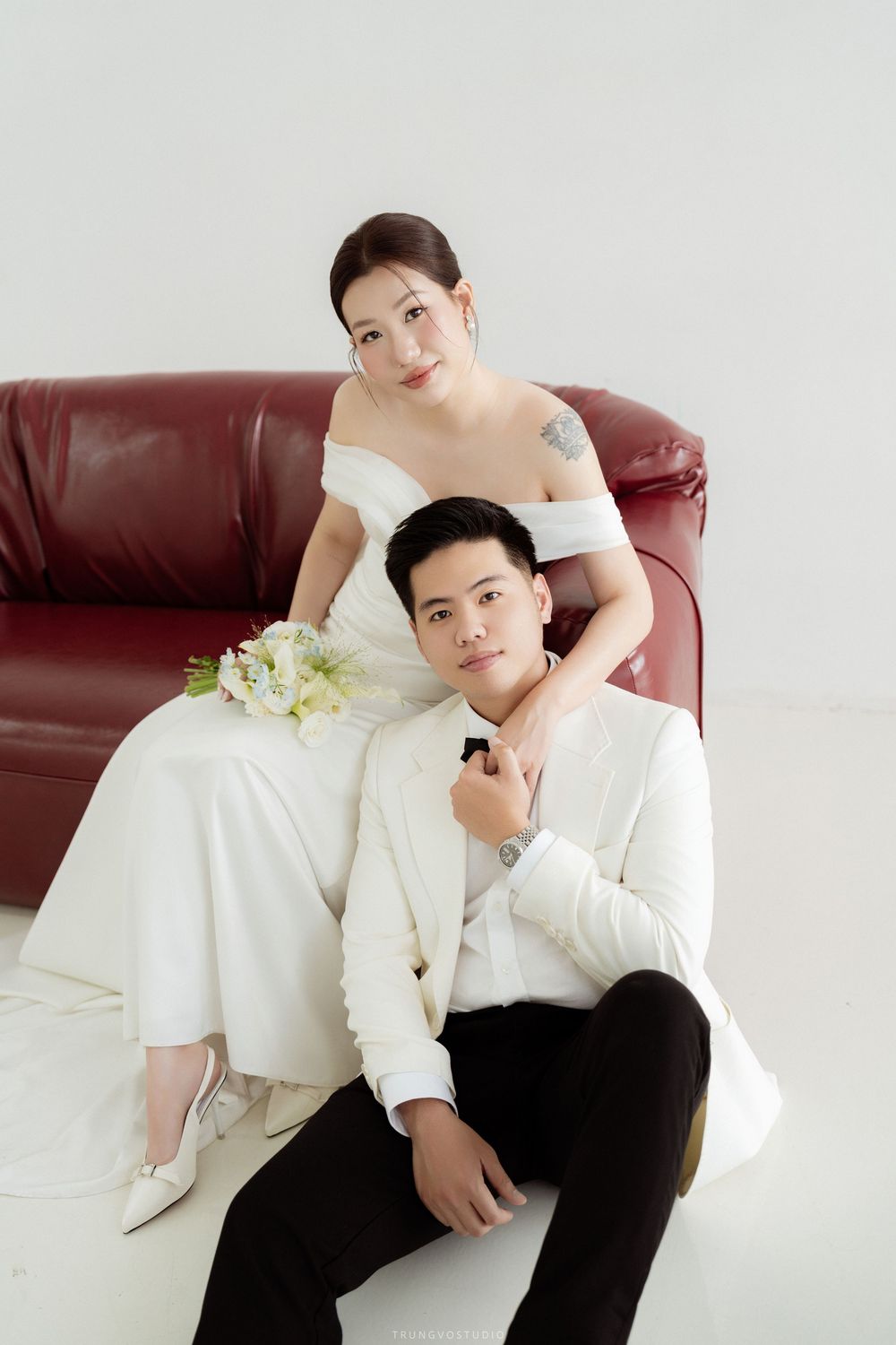Bride in white off-shoulder gown embraces groom seated on floor against red leather sofa in studio.