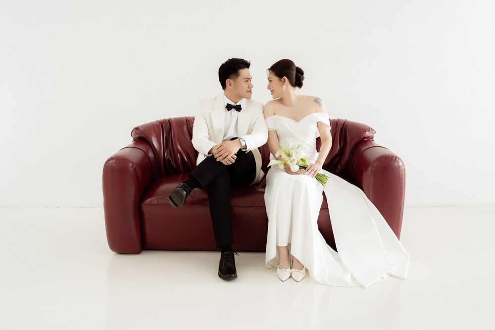 Wedding couple in formal attire sitting together on red leather sofa, smiling at each other lovingly.