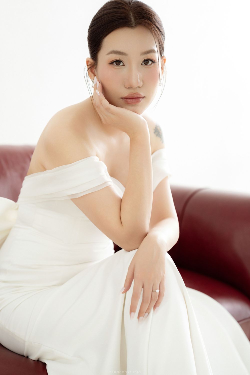 Asian bride in white off-shoulder gown resting chin on hand while seated on red leather sofa.