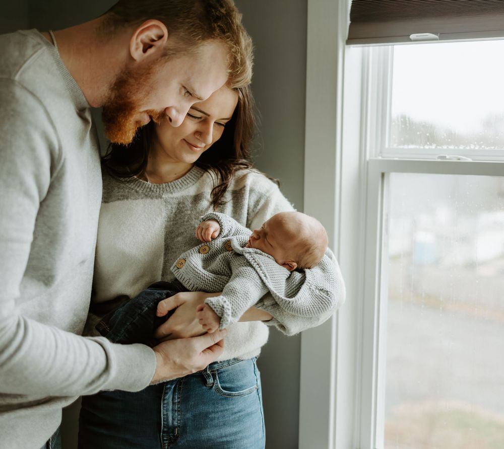 A tender moment shared between a couple holding a newborn baby near a bright window.