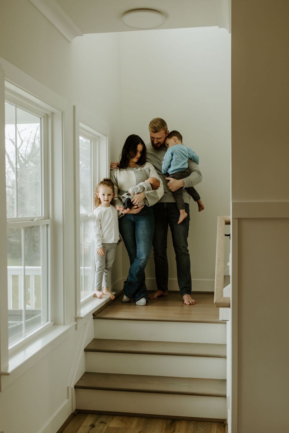 A family stands together on wooden stairs near large windows in their home.