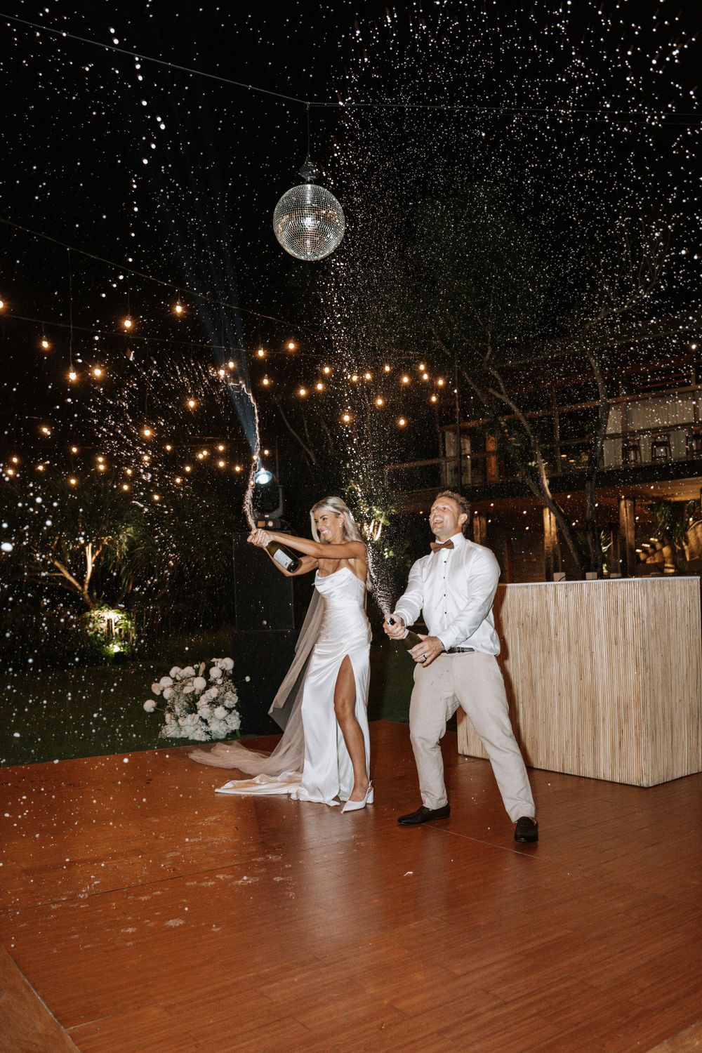 Wedding guests dance on a wooden floor under twinkling lights and a full moon at a nighttime outdoor reception.