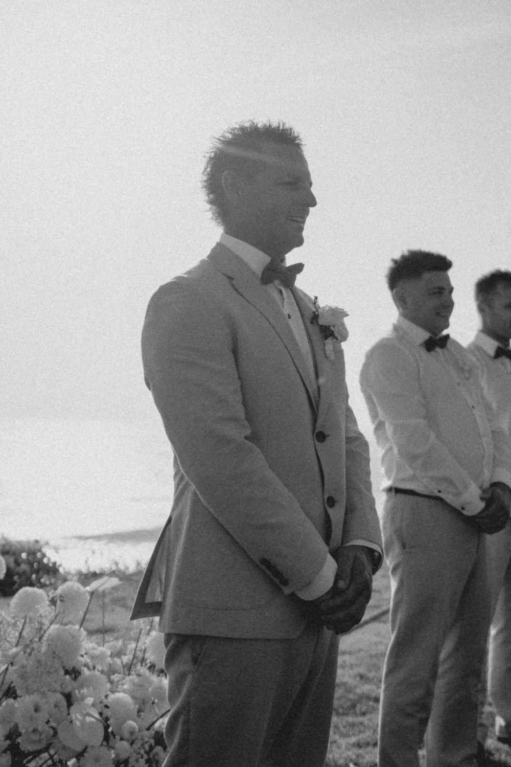 Wedding party members stand in formalwear near ocean beach scenery in black and white photograph.