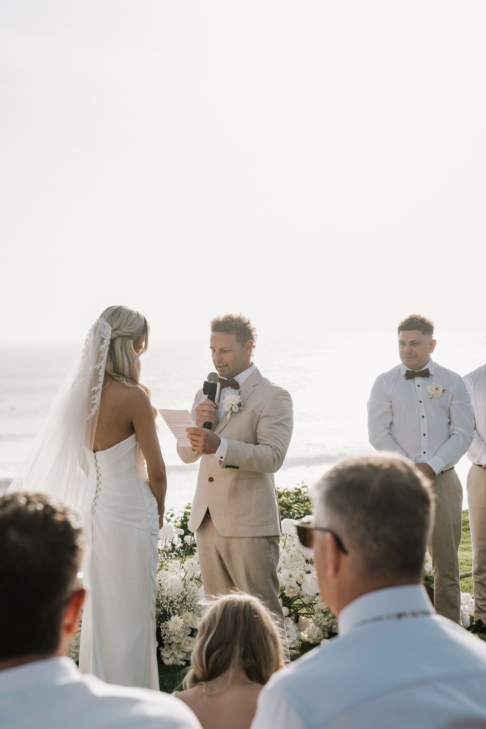 A wedding ceremony takes place outdoors with guests seated watching the couple exchange vows near floral arrangements.