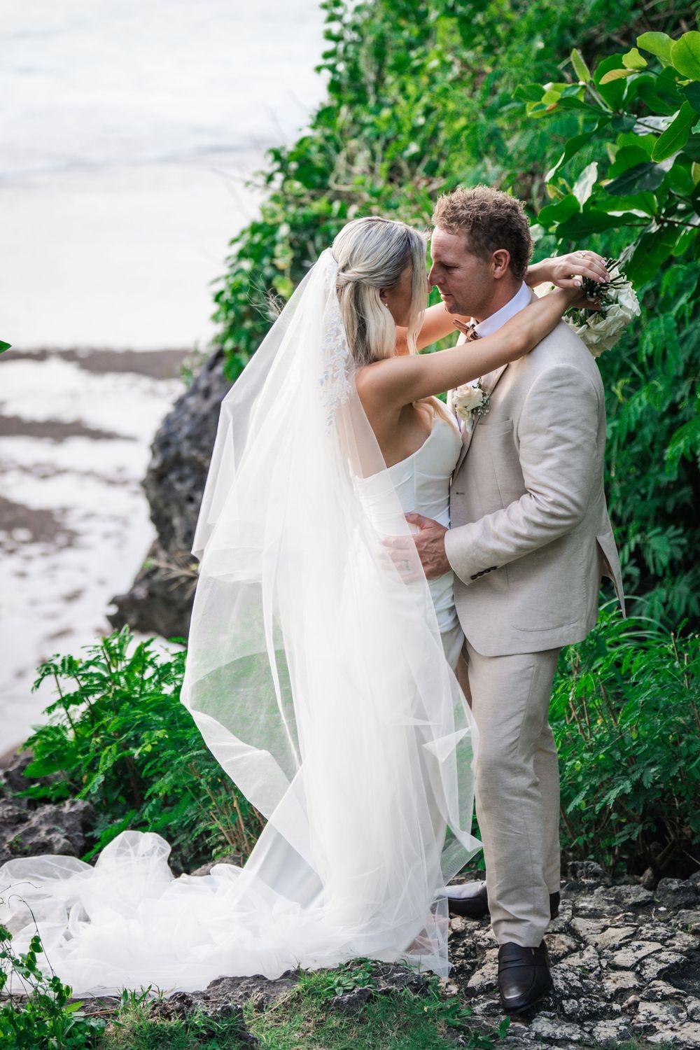 A romantic wedding couple embrace by a waterfront cliff with green foliage in the background, the bride wearing a flowing veil.