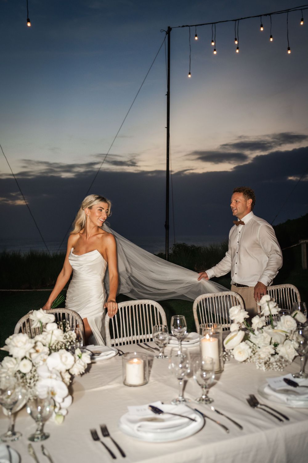 Elegant reception table at outdoor wedding with white floral centerpieces and string lights against dusky sky.