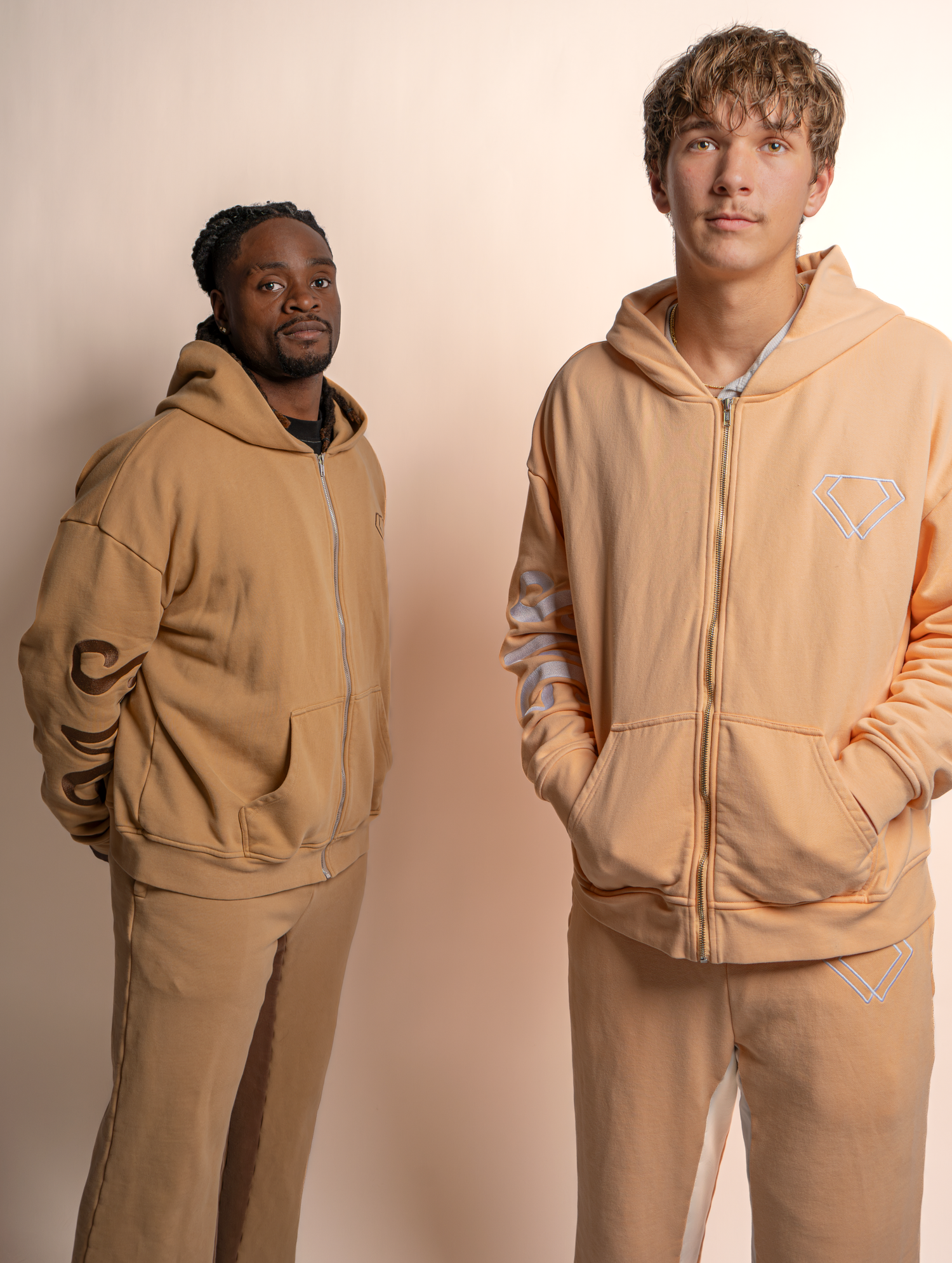 Two people wearing matching beige hooded sweatshirts and sweatpants pose against a light colored background.