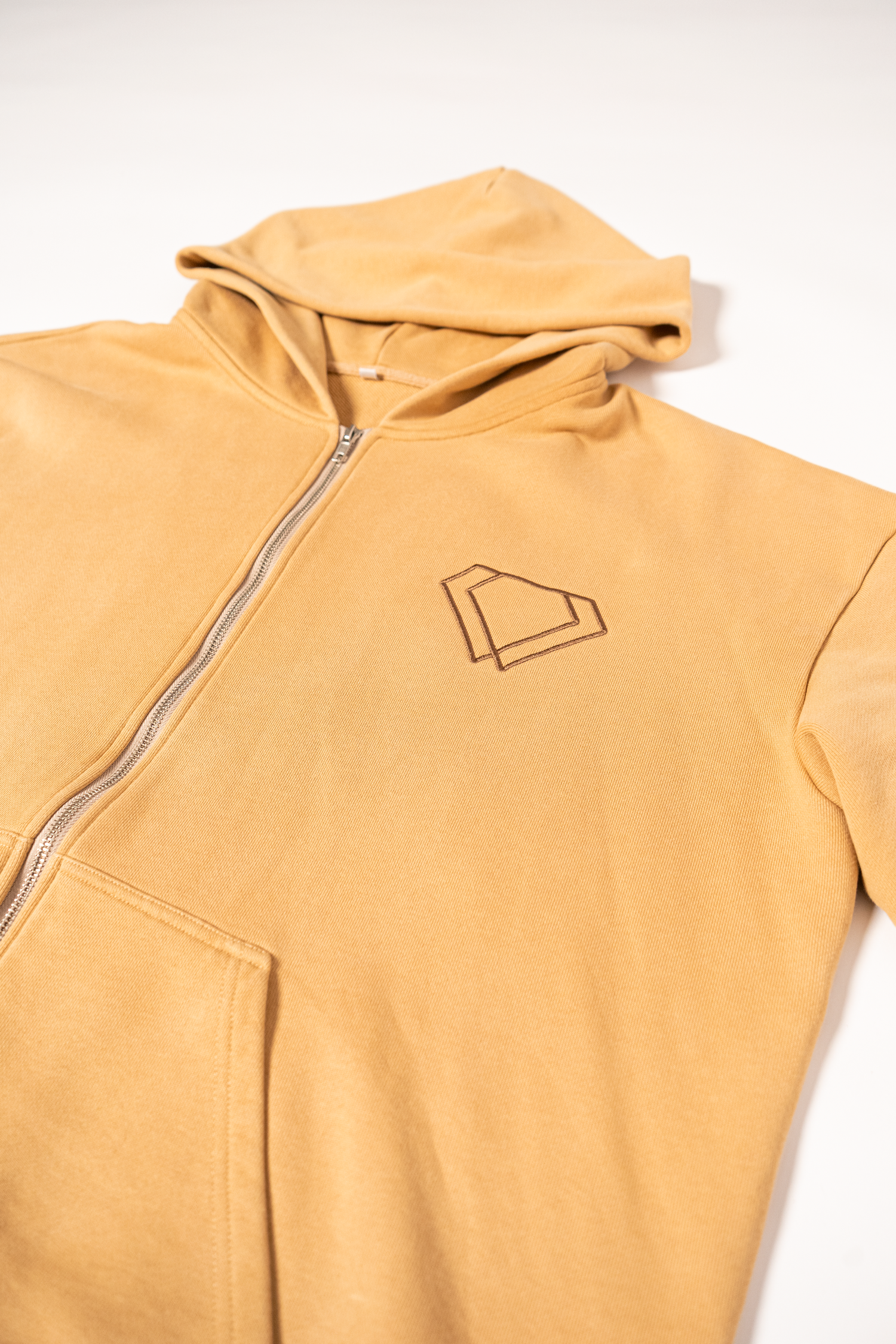 A light beige zip-up hoodie sweatshirt with a minimalist geometric logo design on the chest.