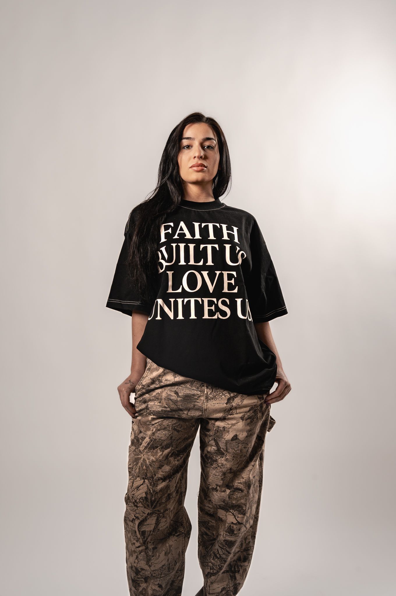 Model wearing black graphic t-shirt with faith message and brown camouflage pants poses against white background.