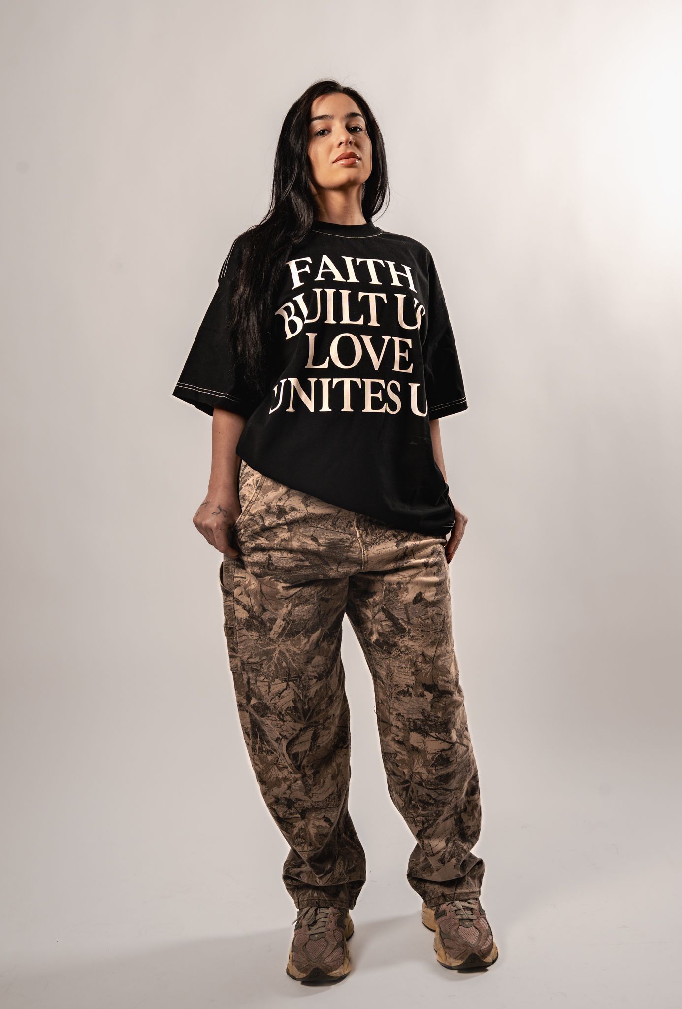 Model in black graphic t-shirt and camo cargo pants posing against white studio background.