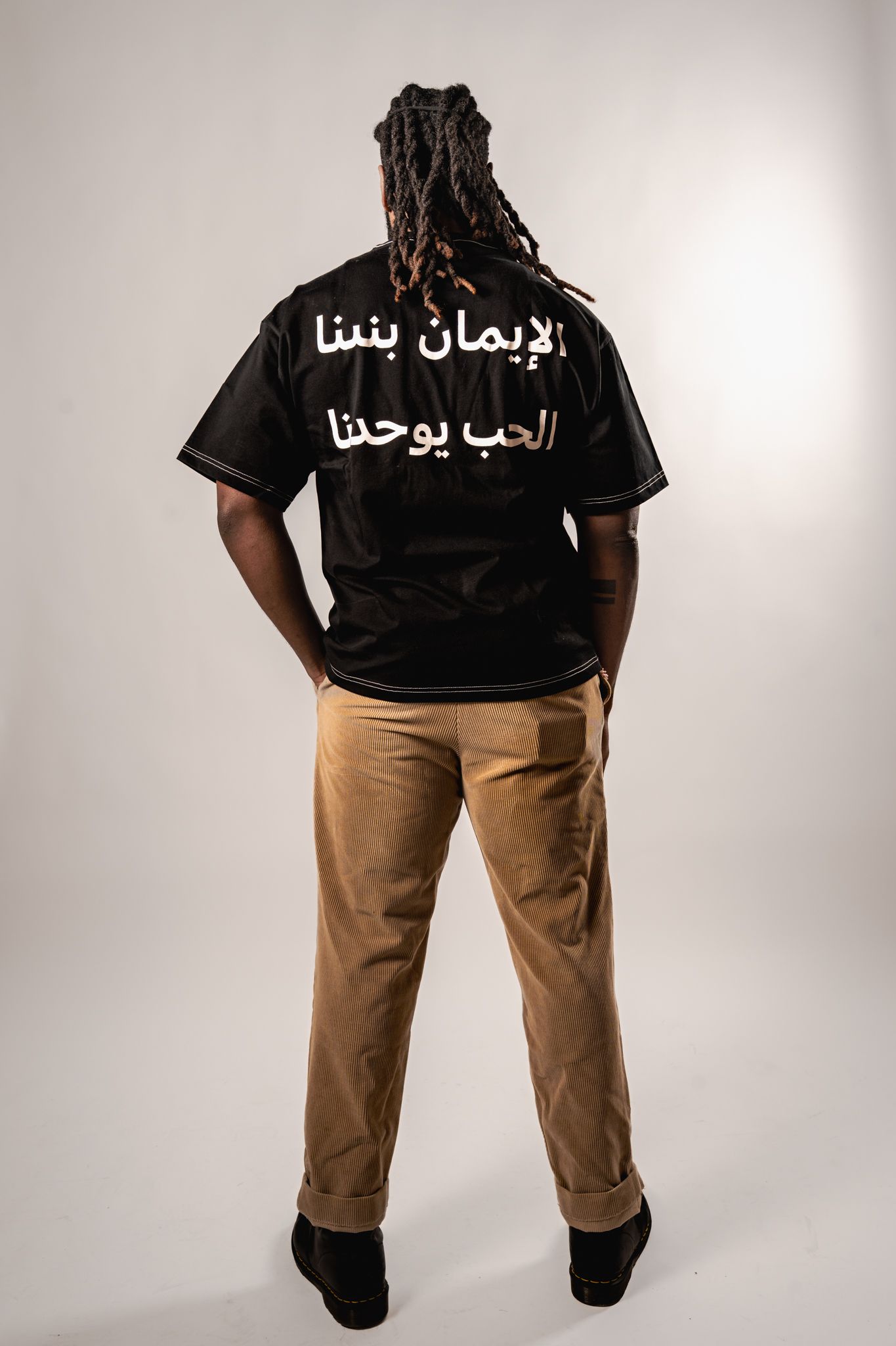 Black t-shirt with Arabic text worn with khaki pants against white backdrop.