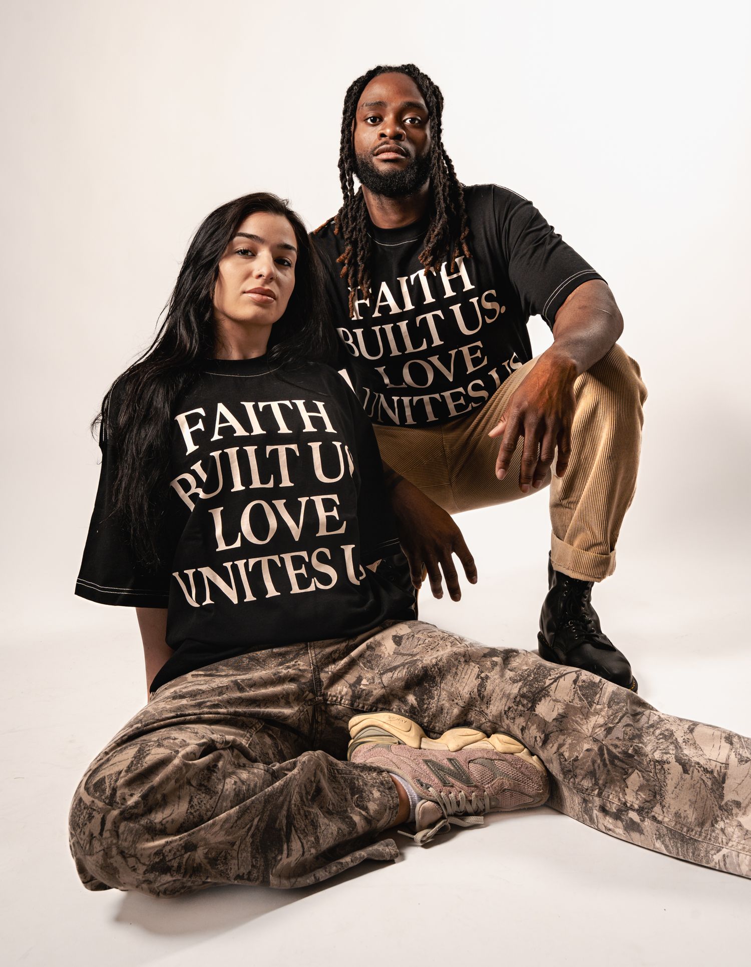 Two people wearing matching black t-shirts with 'FAITH BUILT ON LOVE UNITES' text pose together on rocks against white backdrop.